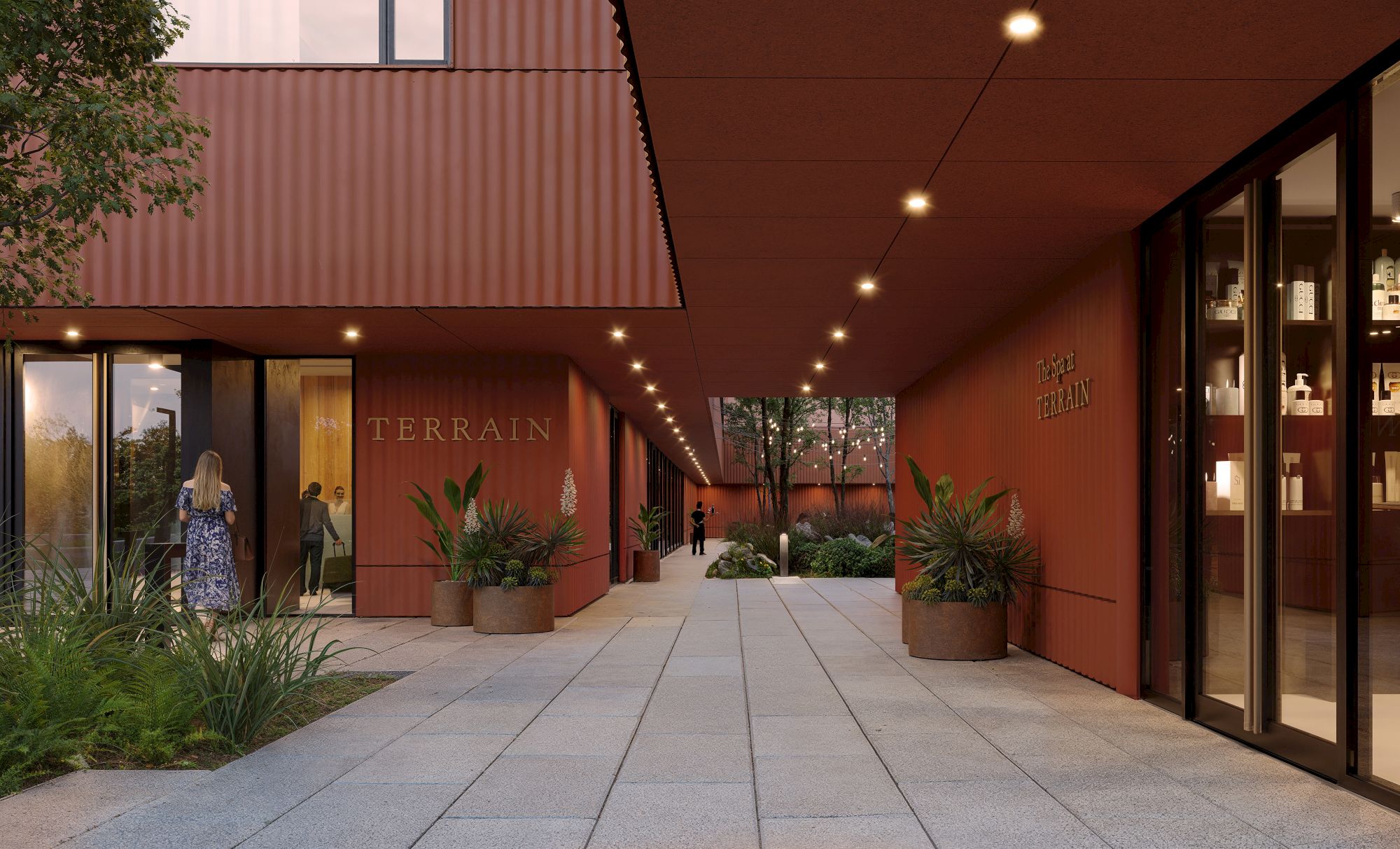 A modern building with red walls, large windows, and plants outside. A person is seen walking near the entrance labeled "Terrain" inside.